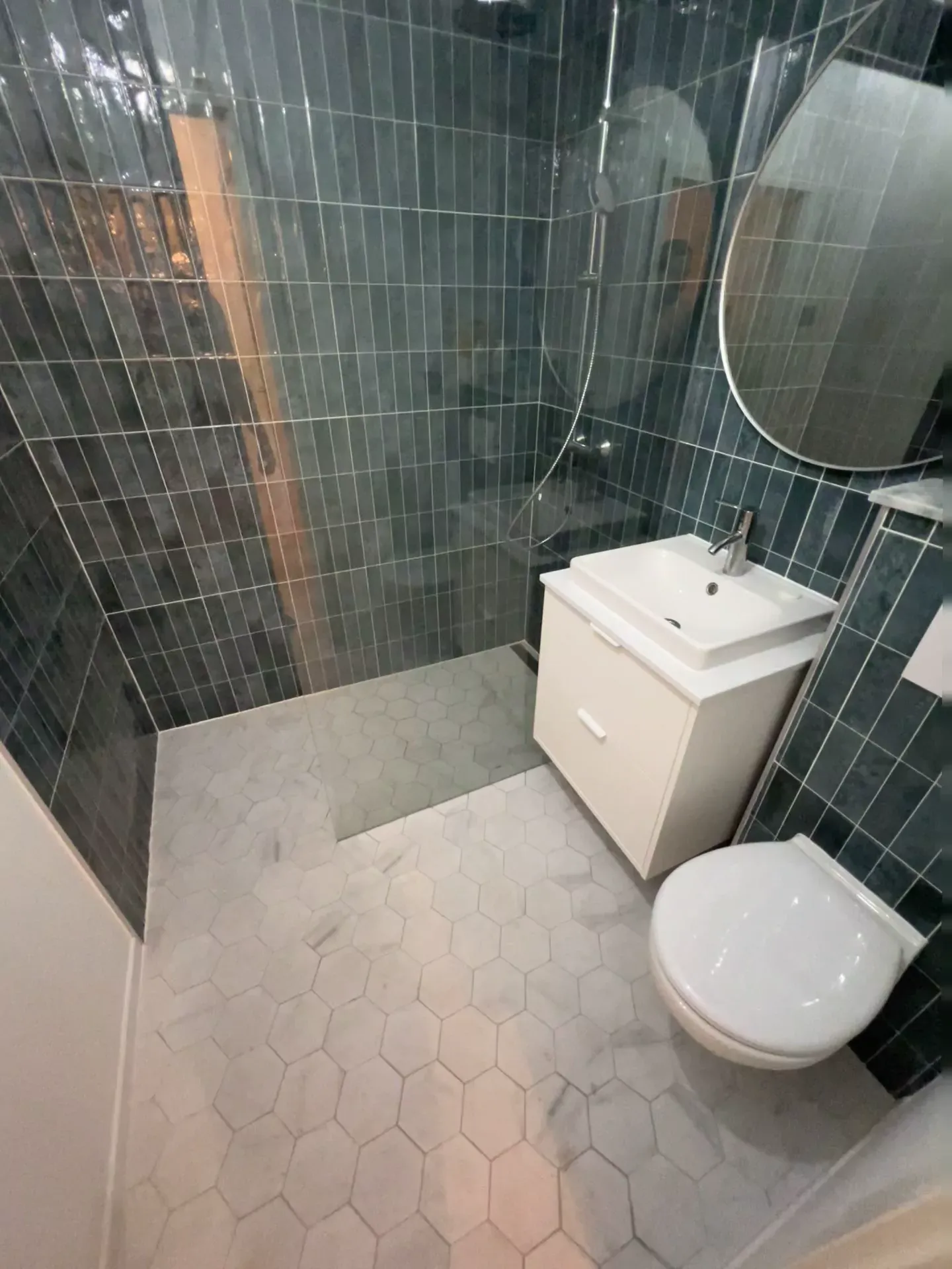 Shower room lined in dark green stack-bond gloss tile, with a walk-in shower behind a curved glass screen, a linear drain, a white wall-hung vanity, a wall-hung WC, a round mirror and a marble-effect hexagon floor.